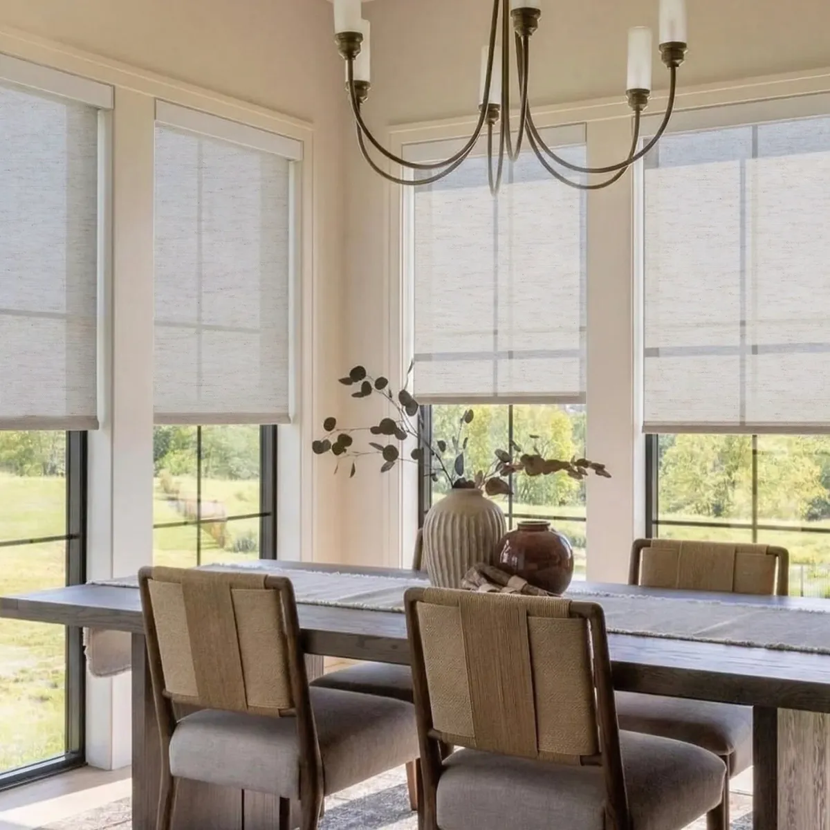 Dining room with light-filtering motorized shades lowered over floor-to-ceiling windows, a wood table and woven chairs beneath a chandelier