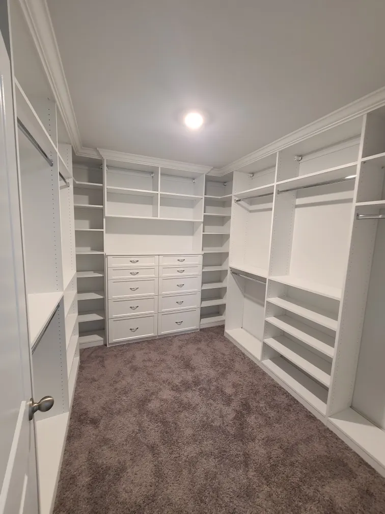 White walk-in closet with a built-in corner dresser flanked by hanging rods and open shelving, over plush carpet