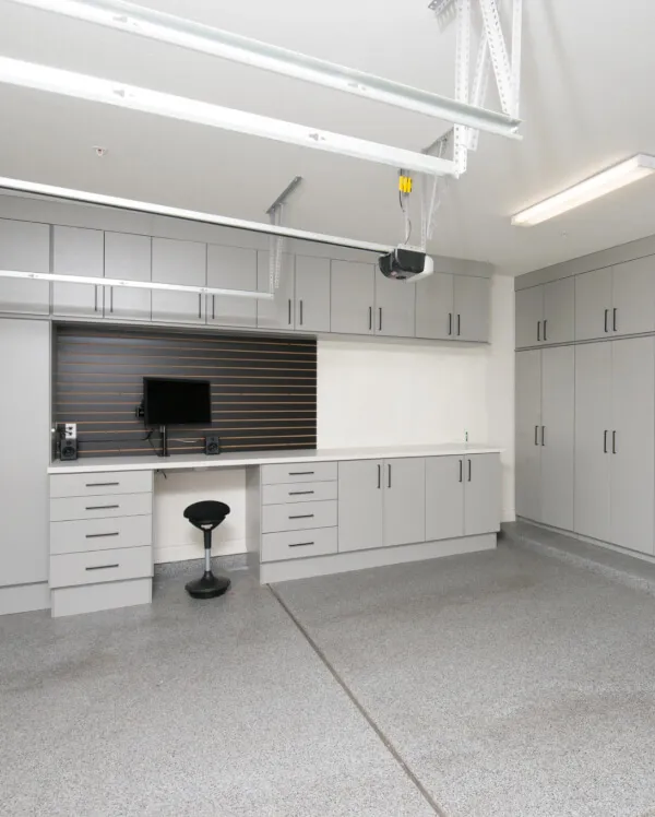 Garage workstation with gray overhead cabinets, a wood-look slatwall backdrop, wall-mounted monitor and a rolling stool over an epoxy floor
