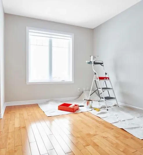 Empty room prepped for painting, with a step ladder, paint tray and drop cloths spread over a hardwood floor
