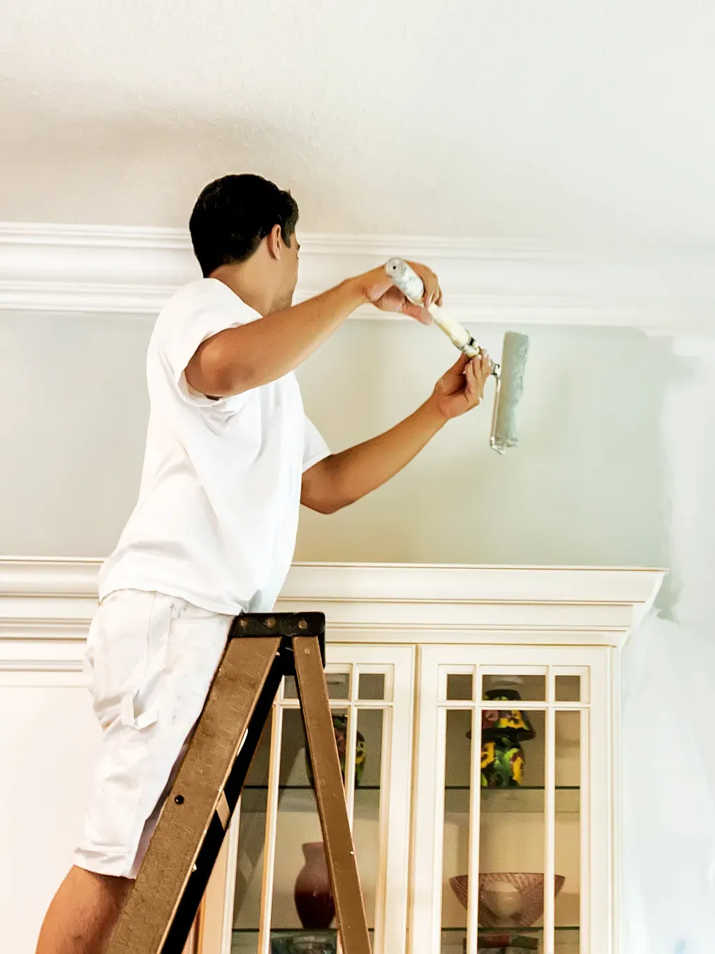 Painter on a ladder rolling fresh paint above a built-in cabinet with crown molding