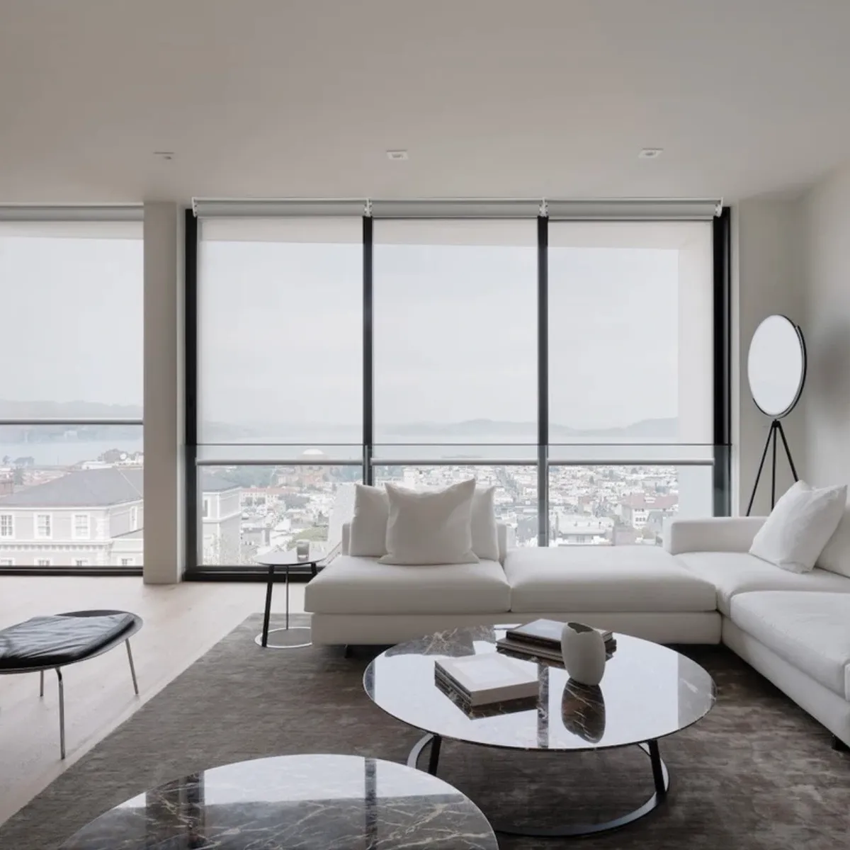 Living room with light-filtering motorized shades lowered across floor-to-ceiling windows overlooking a bay and city view, white sectional sofa and round marble coffee tables